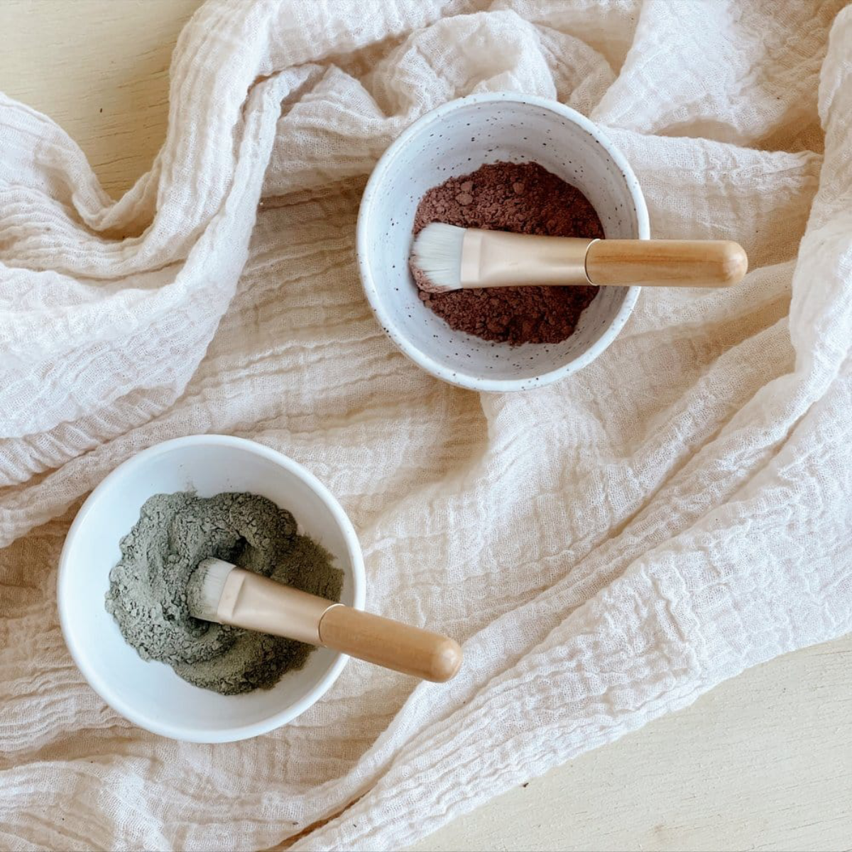 Two bowls with green and brown powders and wooden spoons on a beige fabric background